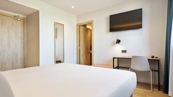 Een hotelkamer met een groot bed, een bureau, een stoel, een aan de muur bevestigde televisie en een deur die naar een andere kamer leidt.