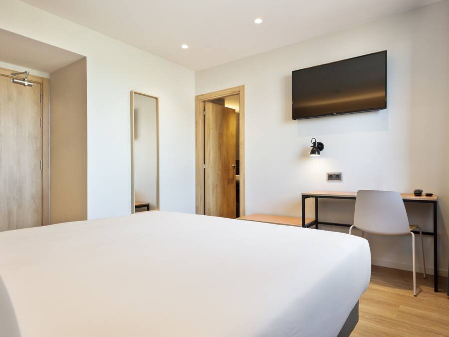 Een hotelkamer met een groot bed, een bureau, een stoel, een aan de muur bevestigde televisie en een deur die naar een andere kamer leidt.