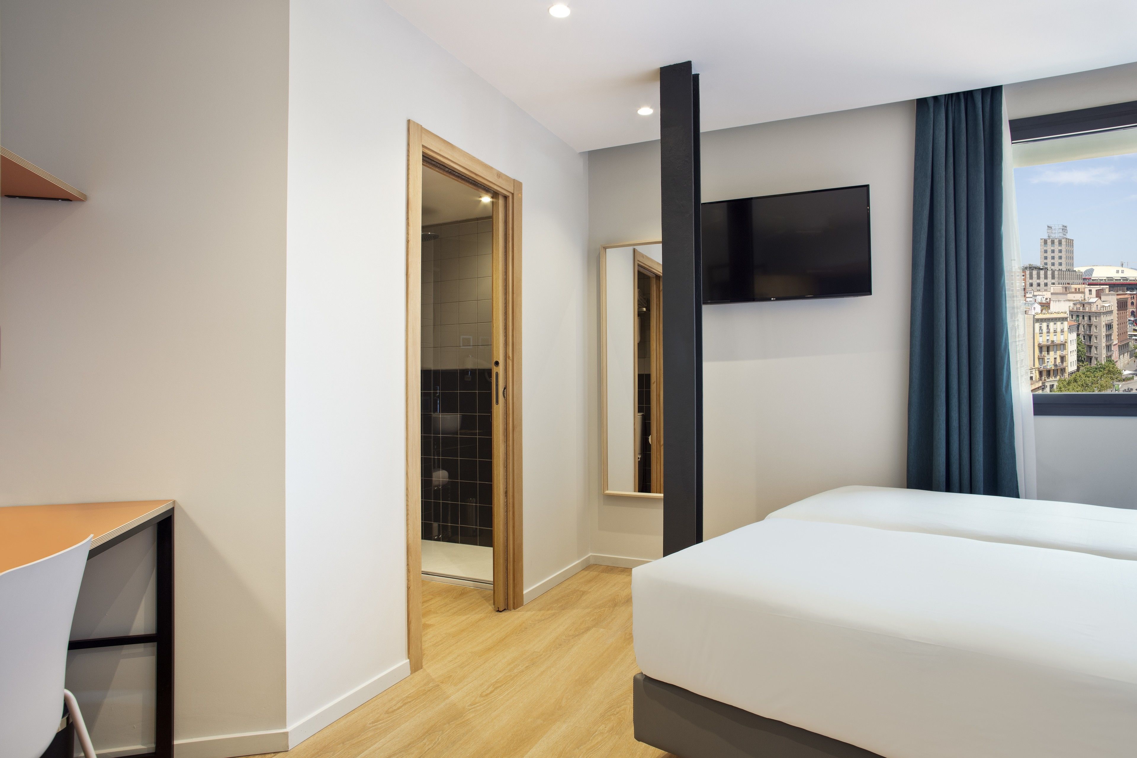 A hotel room with a bed, a desk, a TV mounted on the wall, and a view of the city through the window.