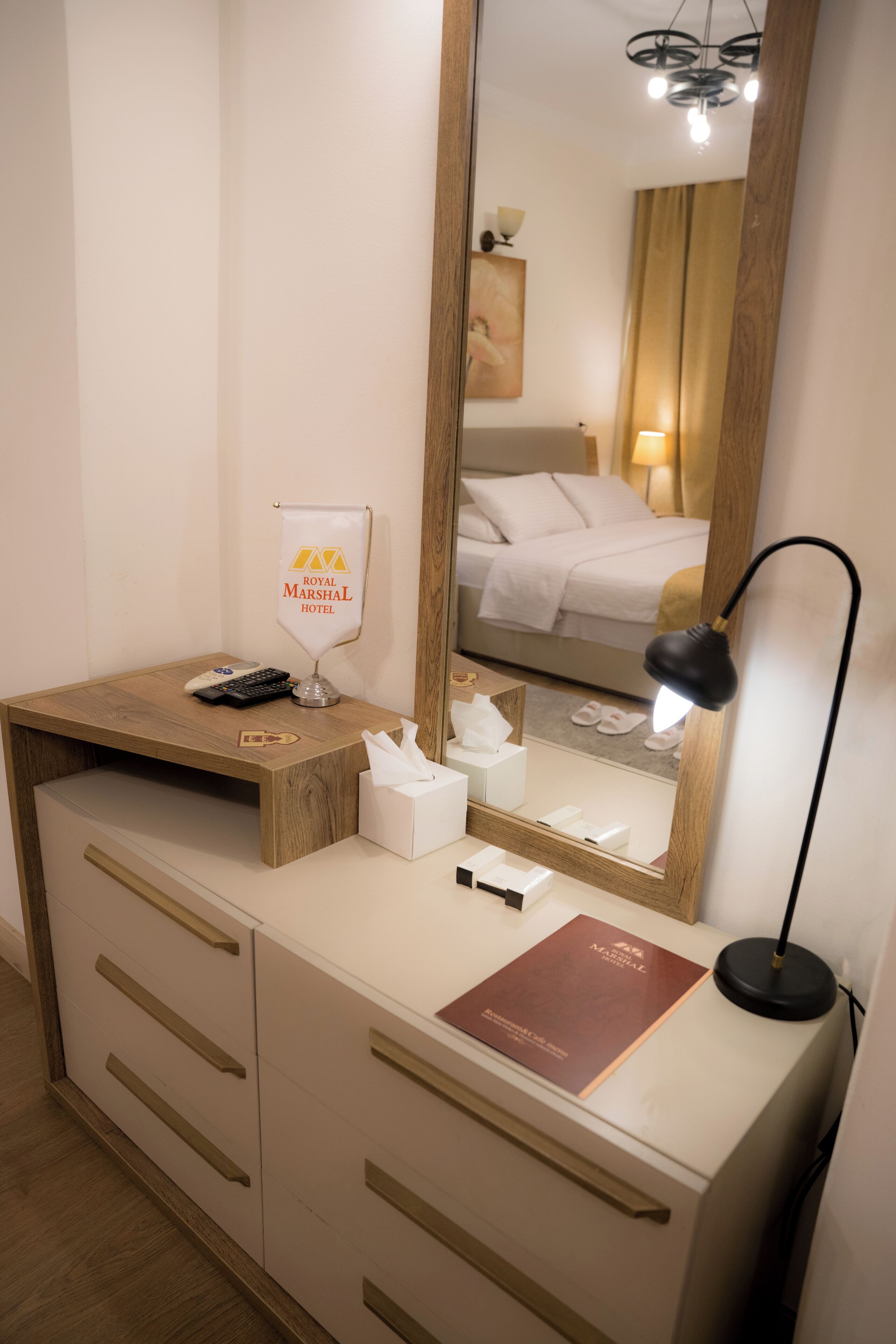 Une chambre d’hôtel avec un bureau en bois, un miroir, une lampe, une boîte de mouchoirs, une télécommande et une brochure.