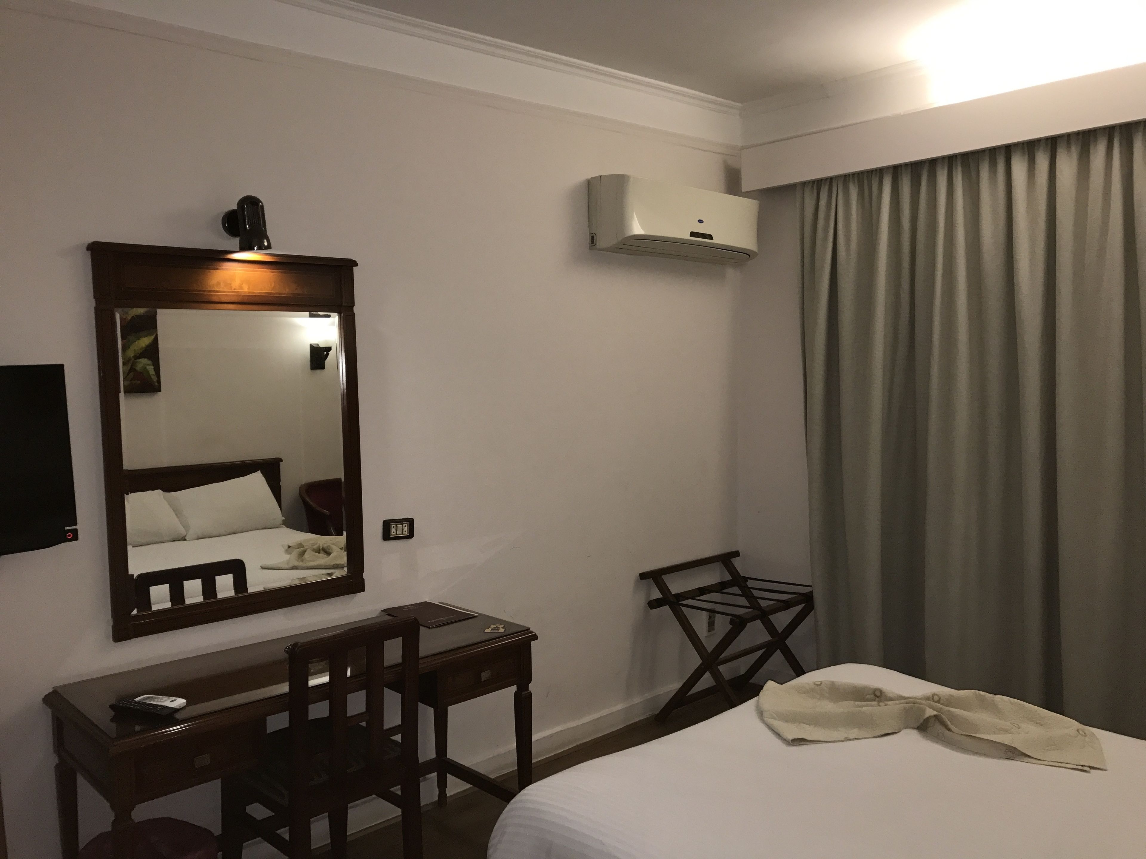 Quarto de hotel com cama, escrivaninha, cadeira, espelho, ar condicionado e janela com cortinas.