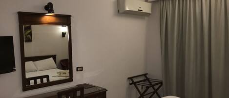 Quarto de hotel com cama, escrivaninha, cadeira, espelho, ar condicionado e janela com cortinas.