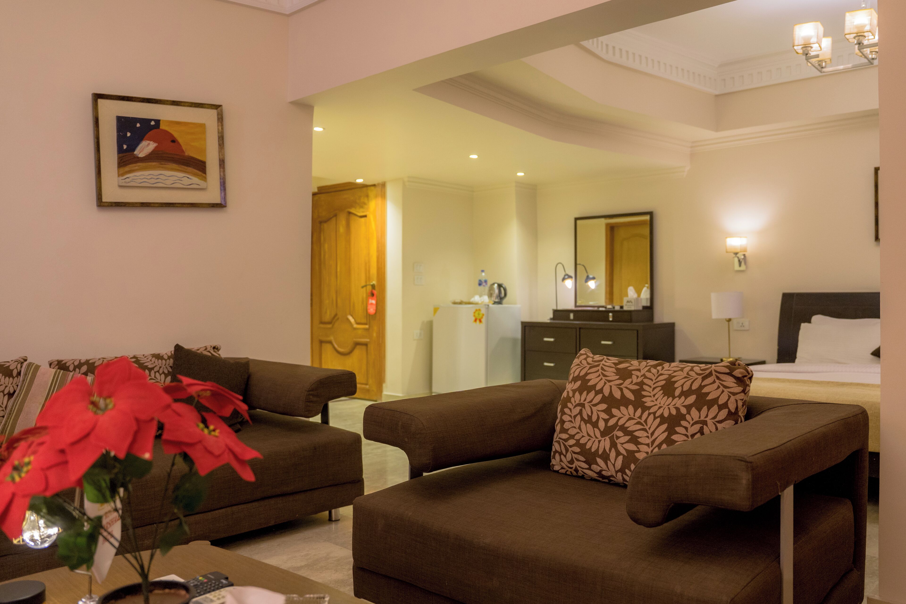 Quarto de hotel com sofá marrom, uma mesinha com um vaso de flores vermelhas, cama com lençóis brancos, cômoda e um espelho.