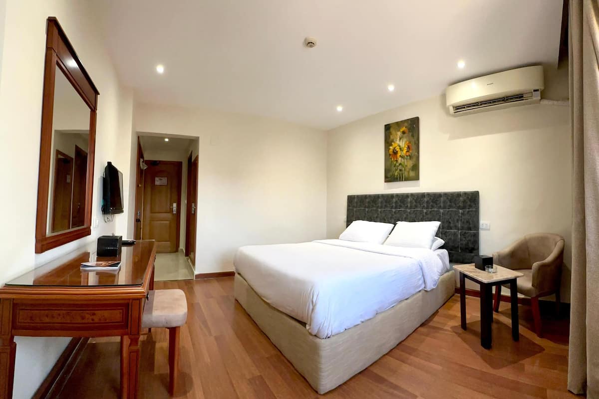 A hotel room with a bed, a desk, a chair, an air conditioning unit, and a painting on the wall.