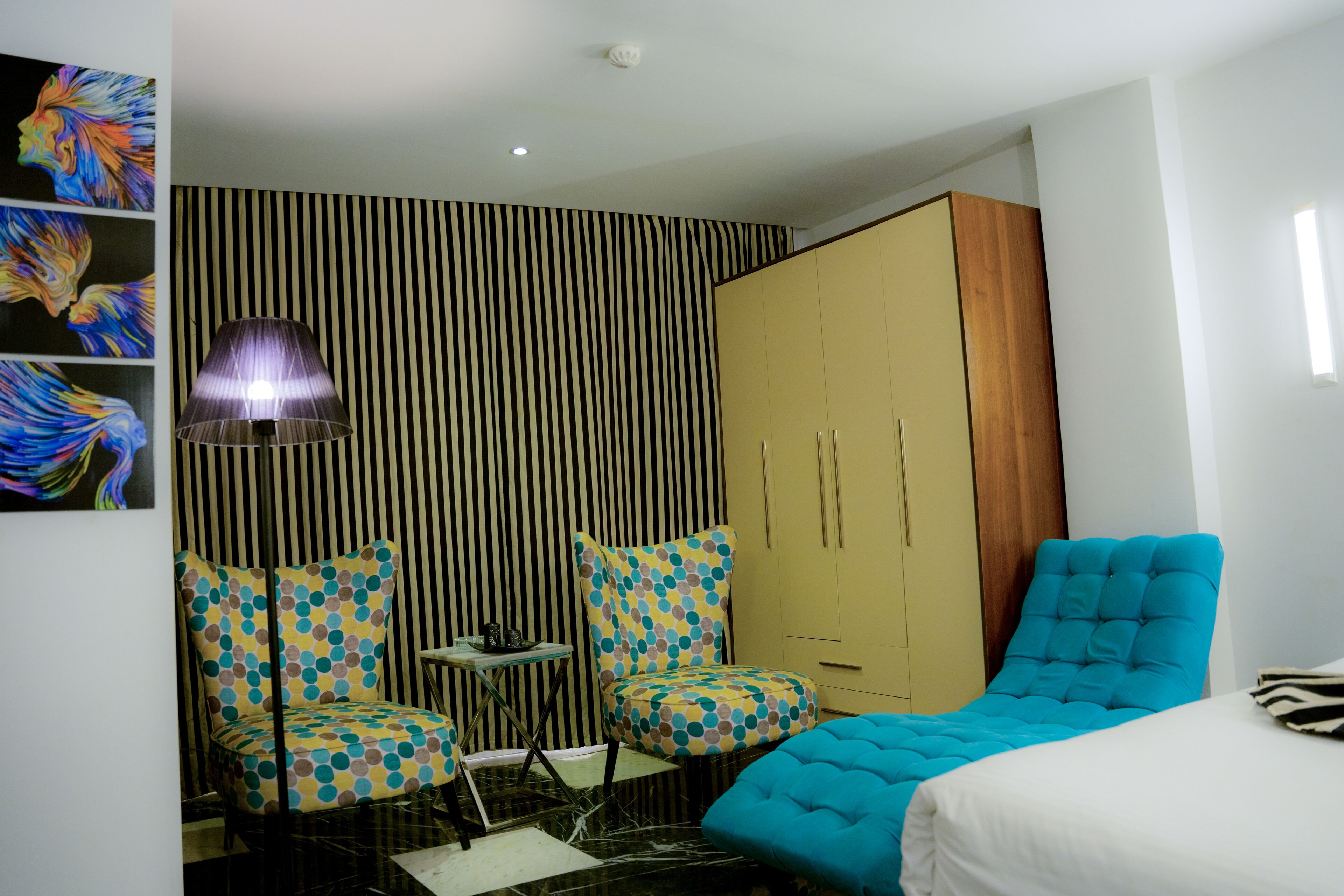 Une chambre moderne avec un papier peint à rayures, un lit doté d’une tête de lit rembourrée en tissu bleu, une armoire et deux chaises à motifs à pois.