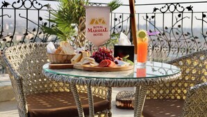 Une table en osier surmontée d’une plaque de verre, une enseigne indiquant l’hôtel Royal Marshal, une variété de fromages, des raisins et un cocktail posés sur la table.