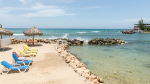 A beachside lounge with sun loungers and a thatched umbrella, a rocky shoreline, and a clear view of the ocean. - Pipers Cove - Runaway Bay (Runaway Bay)