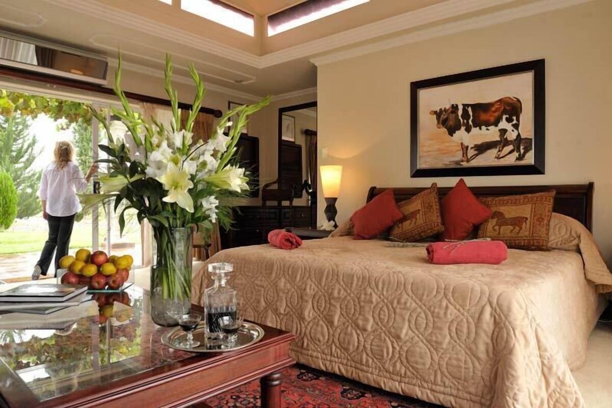 A hotel room with a large bed, a vase of flowers, a tray with fruits and a bottle, and a framed picture on the wall.