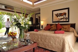Quarto de hotel com uma cama grande, um vaso de flores, uma bandeja com frutas e uma garrafa, e um quadro na parede.