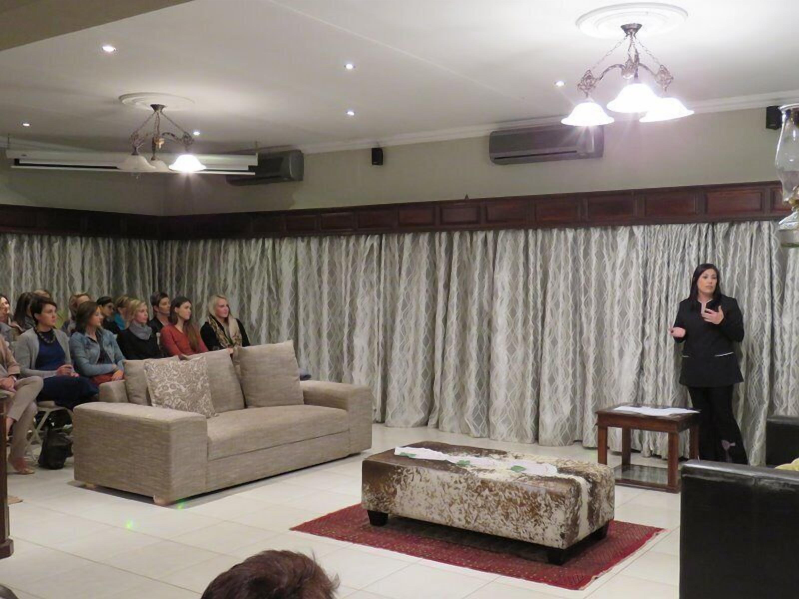 A room with a sofa, ottoman, and a standing speaker addressing an audience.