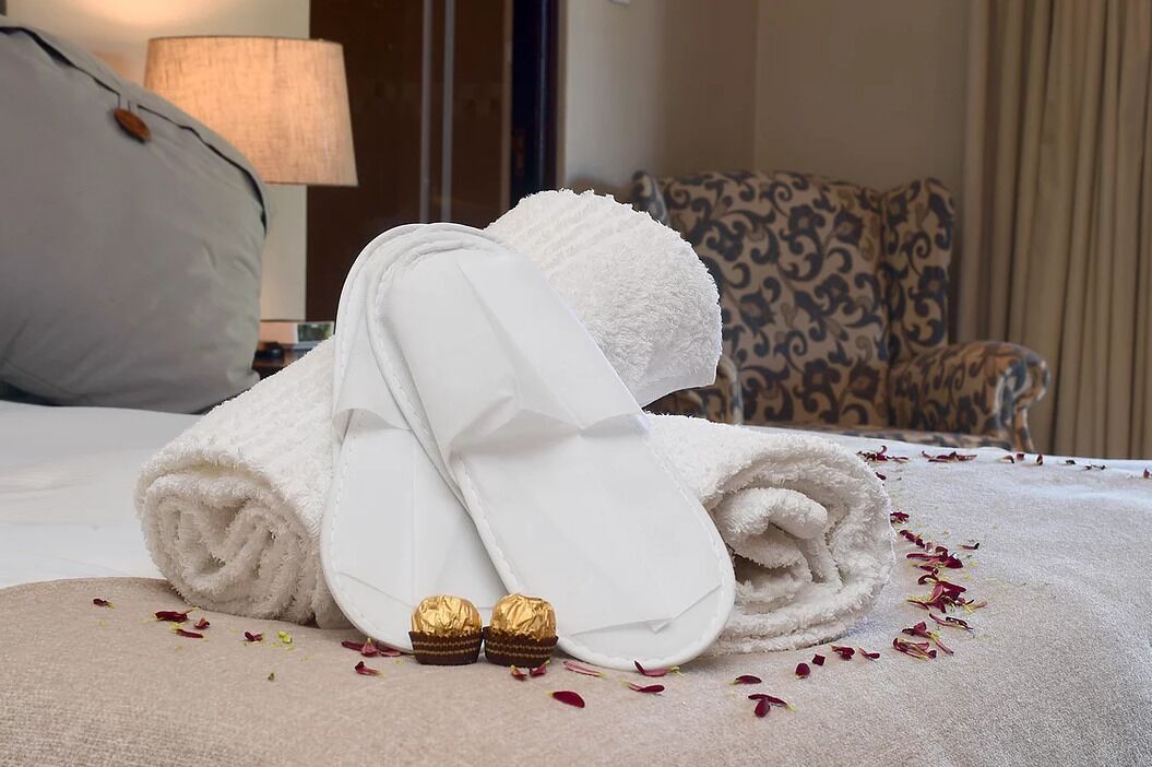 A neatly made bed with rolled white towels and two golden chocolates on a beige bedspread, with a patterned armchair and a lamp in the background.