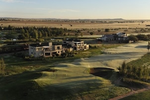 Um campo de golfe com vários edifícios, incluindo uma casa grande e uma construção menor, rodeado por campos abertos e árvores esparsas.