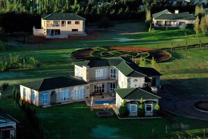 Uma casa grande, com vários andares, e uma piscina, cercada por jardins bem cuidados e um campo de golfe.