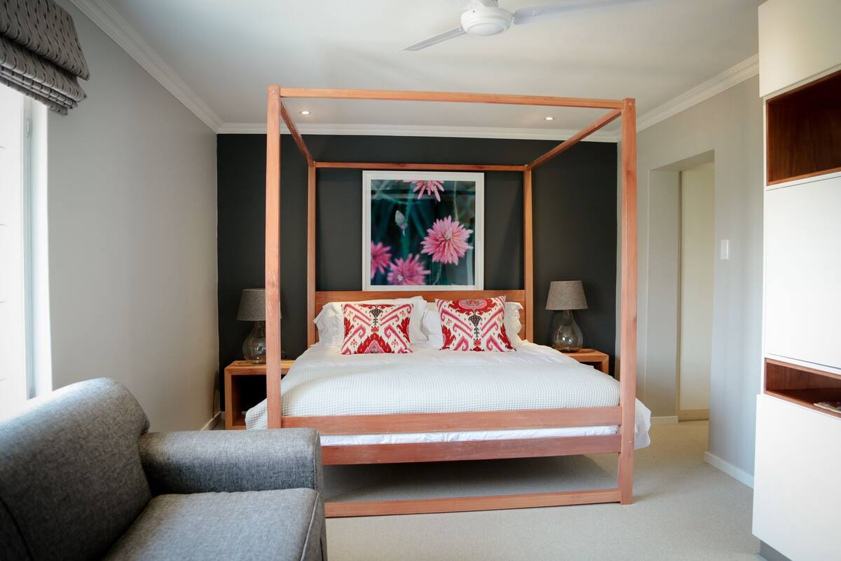 Une chambre à coucher avec un lit à baldaquin, un canapé gris, deux tables de chevet avec des lampes et une grande fenêtre donnant sur un jardin fleuri.