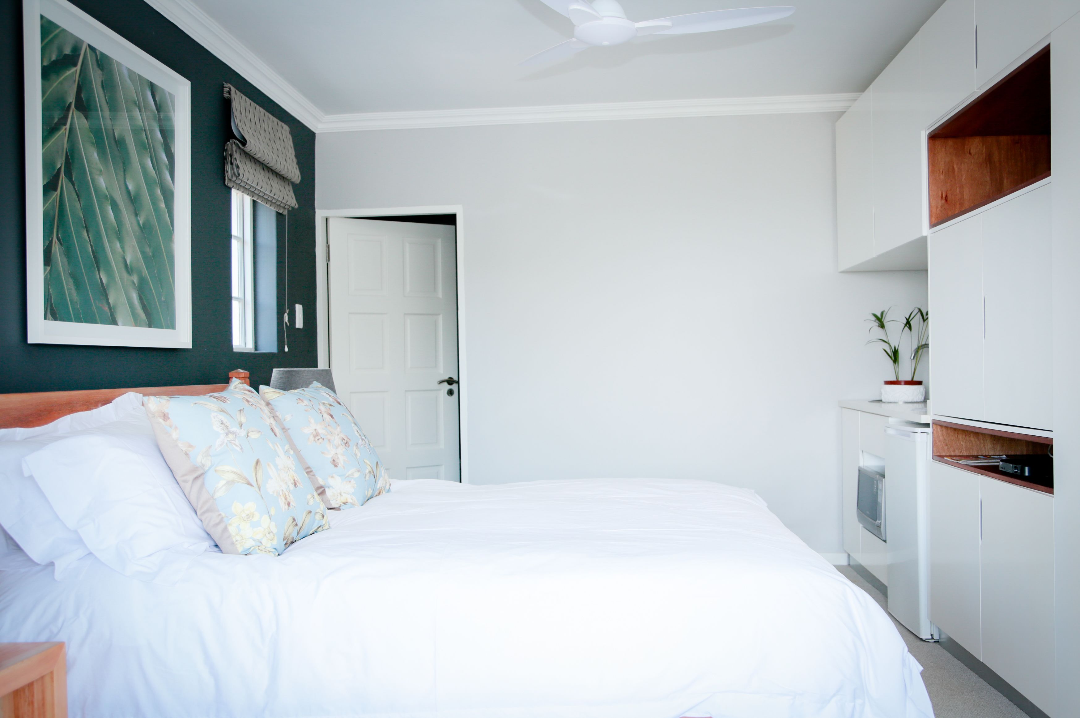 A bedroom with a bed, a ceiling fan, a window with blinds, a door, and a small kitchenette area.