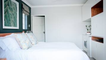 Un dormitorio con cama, ventilador de techo, ventana con persianas, puerta y una pequeña zona de kitchenette.