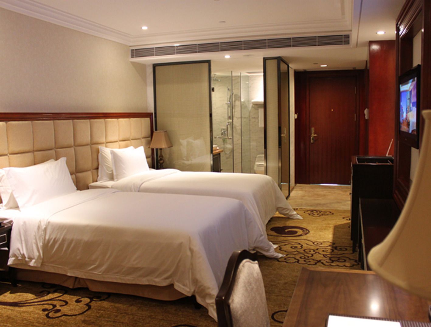 A hotel room with two beds, a TV, a desk, and a bathroom visible through a glass door.