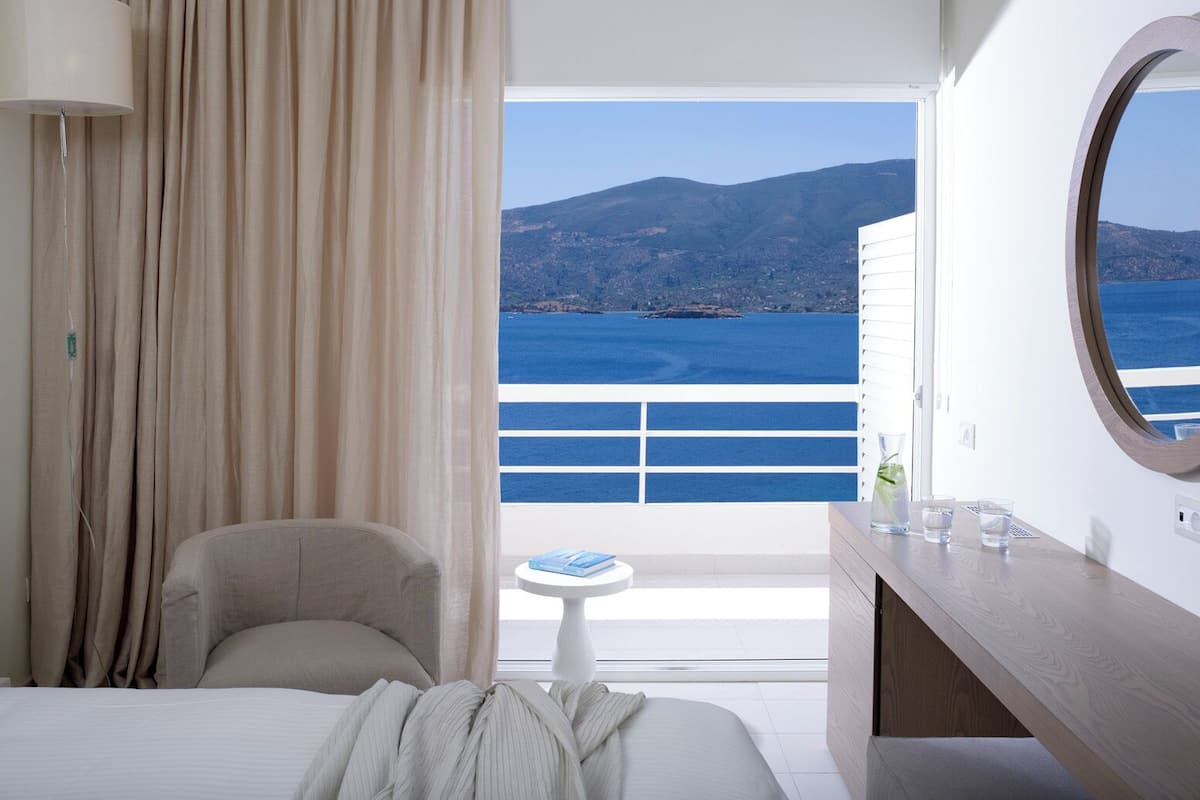 Una habitación de hotel con una cama, una silla, una mesita con vasos, una lámpara y vistas al mar y a las montañas.