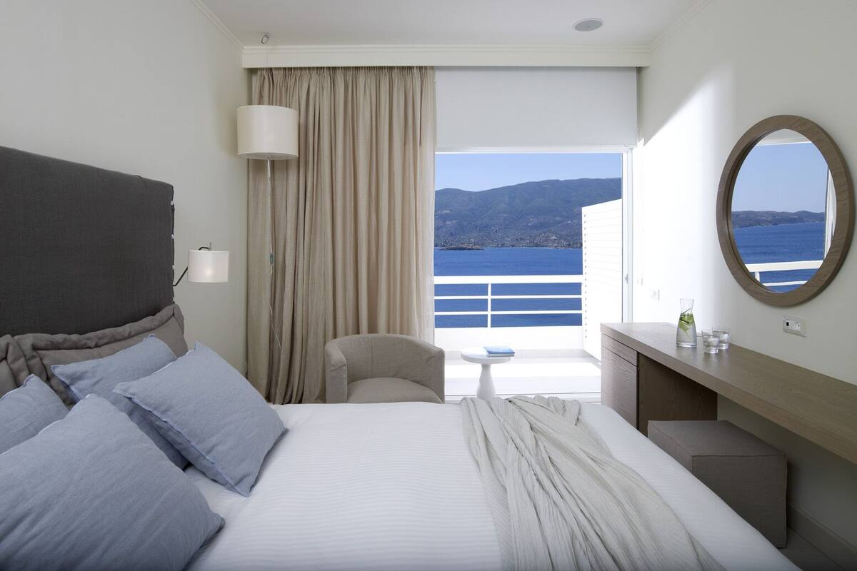 A hotel room with a bed, a chair, a desk, a lamp, and a view of the sea and mountains.