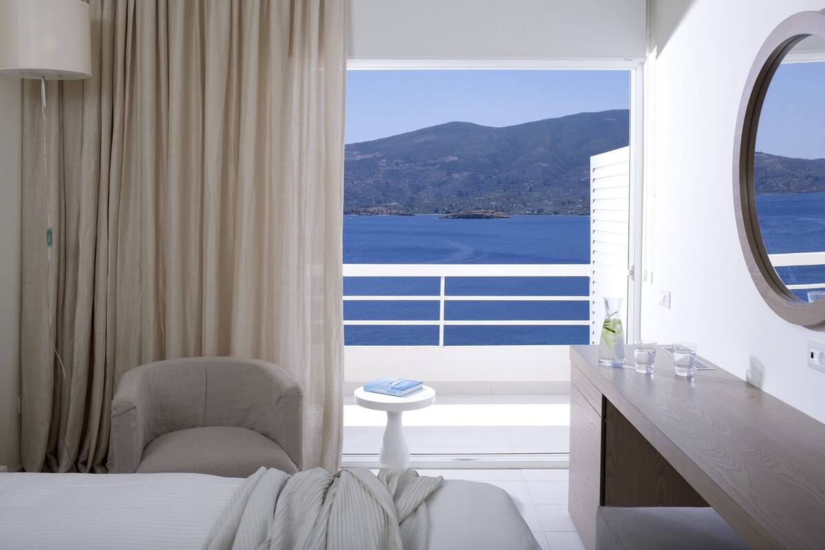 A hotel room with a bed, a chair, a small table, a mirror, and a view of the sea and mountains.