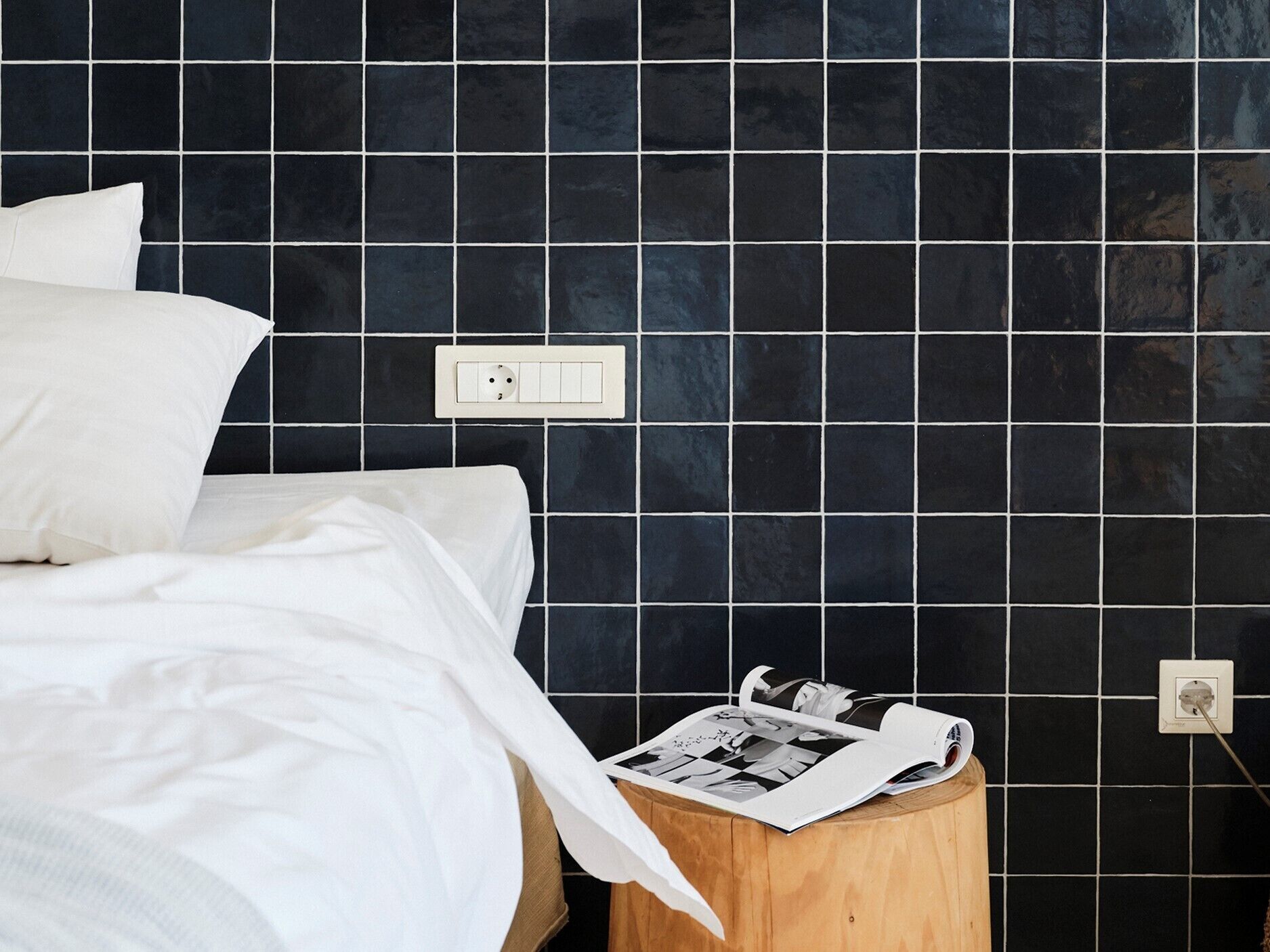 Un dormitorio con cama, ropa de cama blanca, una mesita de noche de madera con una revista y una pared con azulejos negros y enchufes.