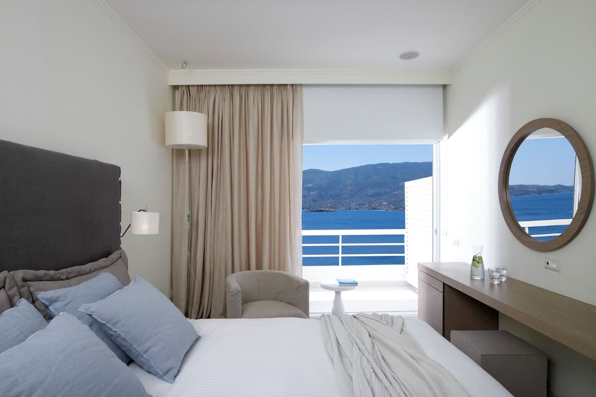 Habitación de hotel con cama, una silla, escritorio, lámpara, espejo y vista al mar.