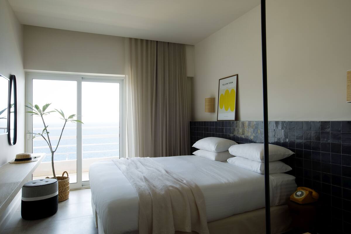 A hotel room with a bed, a plant, a window, and a telephone.