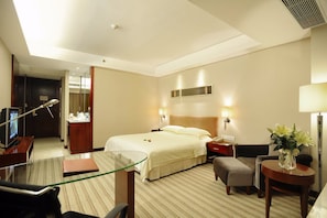 Habitació d'hotel amb llit, taula de nit, làmpada, cadira, escriptori, televisió i una separació de vidre.