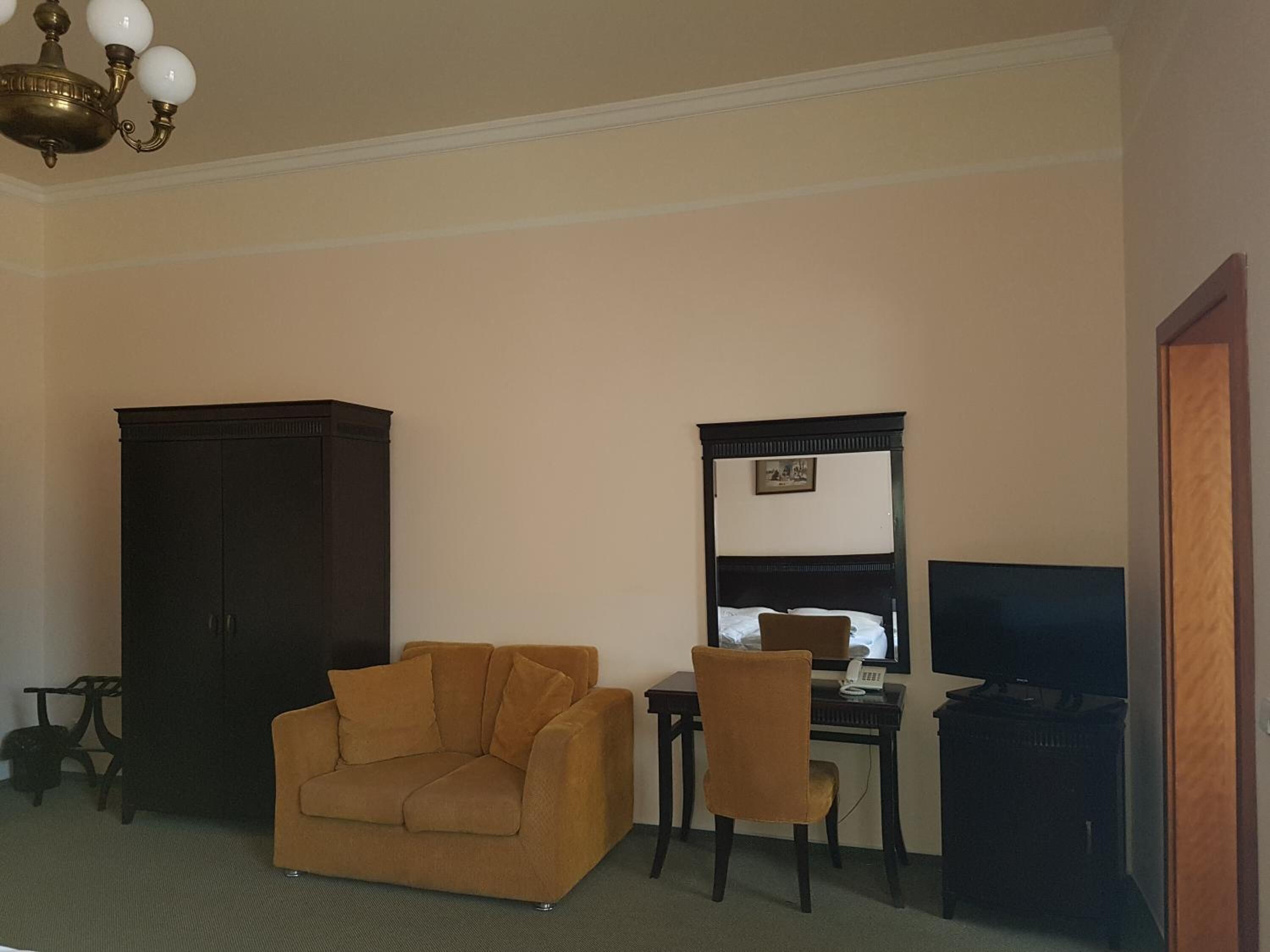 Ein Zimmer mit großem dunklen Holzschrank, einem braunen Sofa, einem kleinen Tisch mit Stuhl, einem Schreibtisch mit Stuhl, einem Fernseher und einer Deckenleuchte.