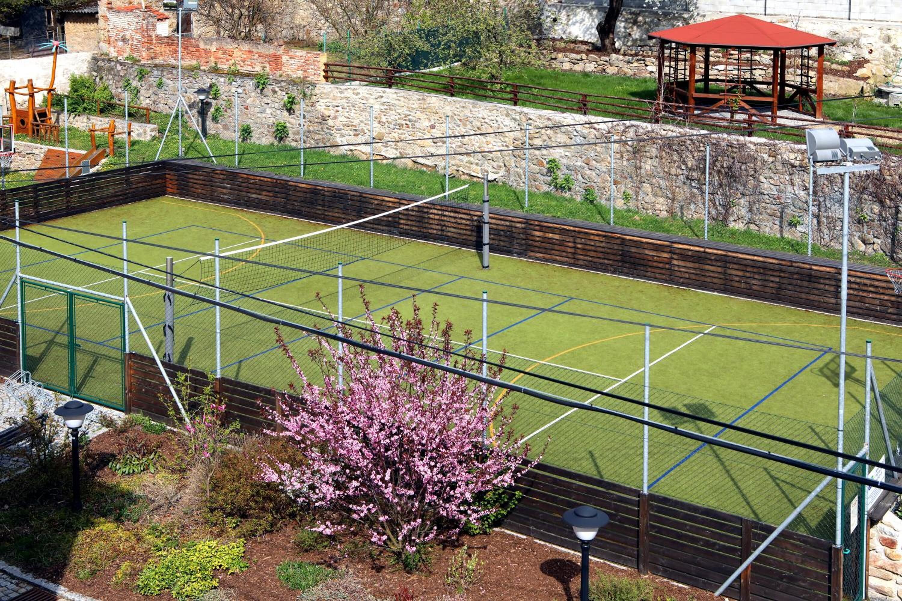 Una cancha de tenis con valla perimetral, un pabellón y un muro de piedra al fondo.