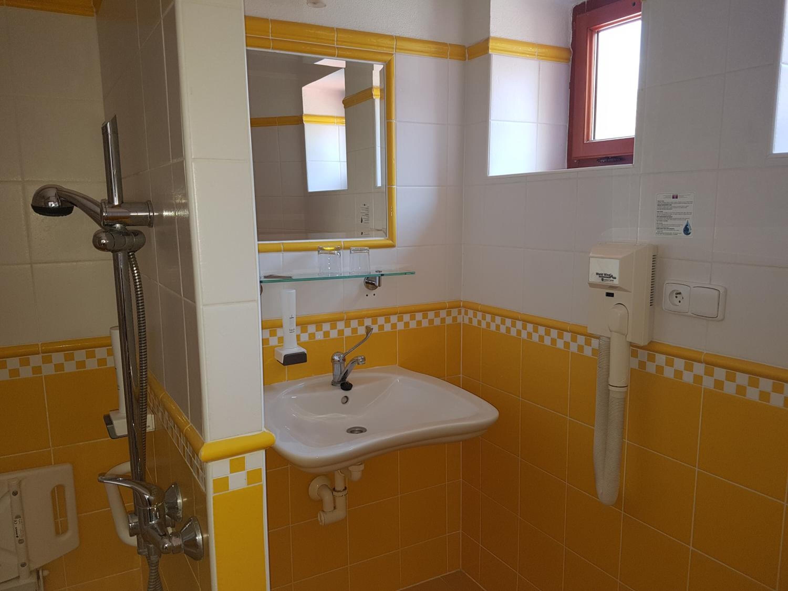 Un baño con baldosas amarillas y blancas, un estante de vidrio, un lavabo, un espejo y una ducha.