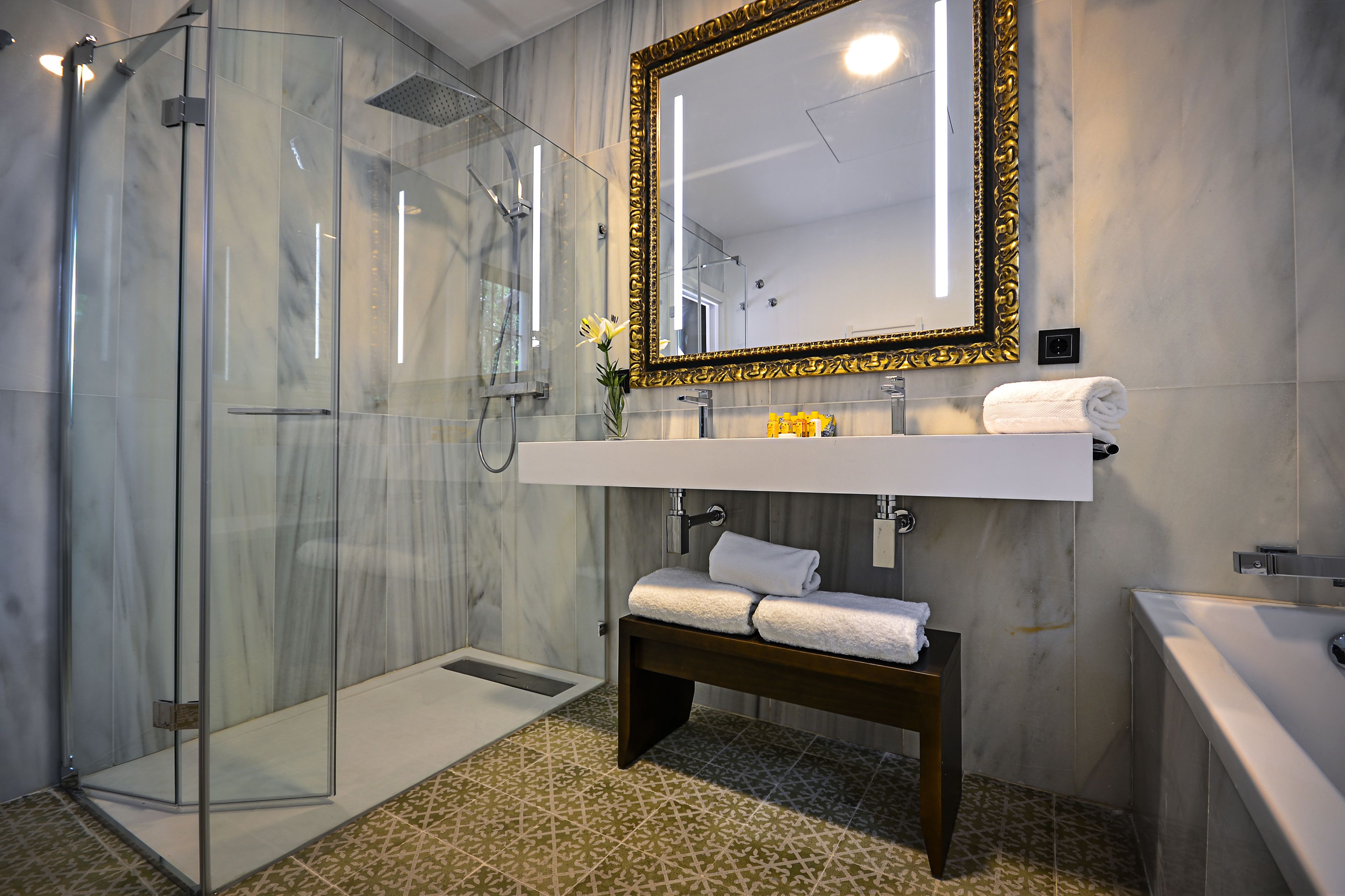 Un baño moderno con cabina de ducha de vidrio, suelo de baldosas de mármol, lavamanos blanco con espejo de marco dorado y un banco con toallas.