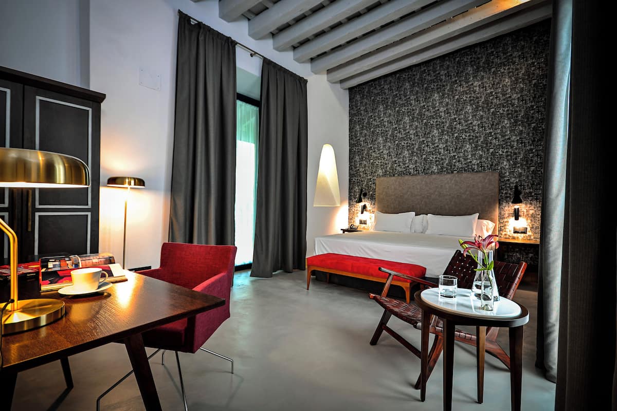 A hotel room with a bed, bedside table, lamp, chair, and a small table with a vase.