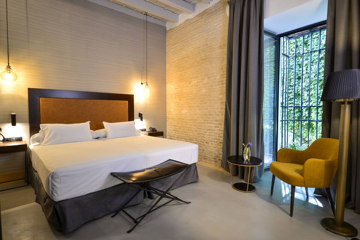 A hotel room with a large bed, a small table, a chair, and a view of greenery through a window.