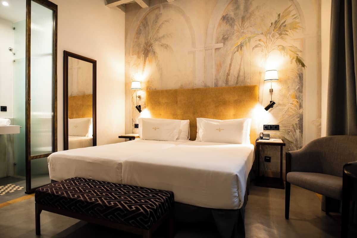 A hotel room with a large bed, two bedside lamps, a bench, a chair, a nightstand, and a wall mural.