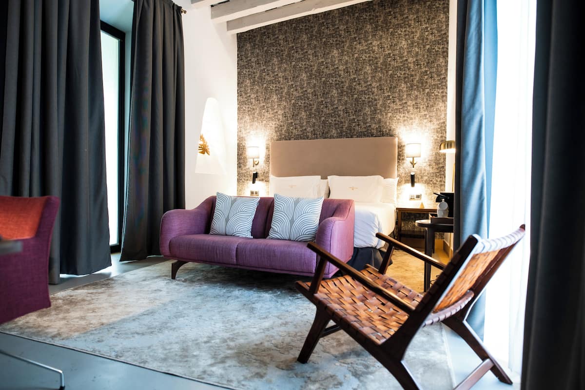 A hotel room with a purple sofa, a wooden chair, a large bed with a headboard, two bedside lamps, and a textured wall.
