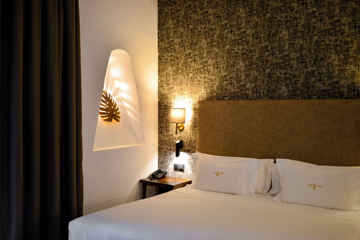 A hotel room with a bed, a nightstand, a lamp, and a wall-mounted light fixture.