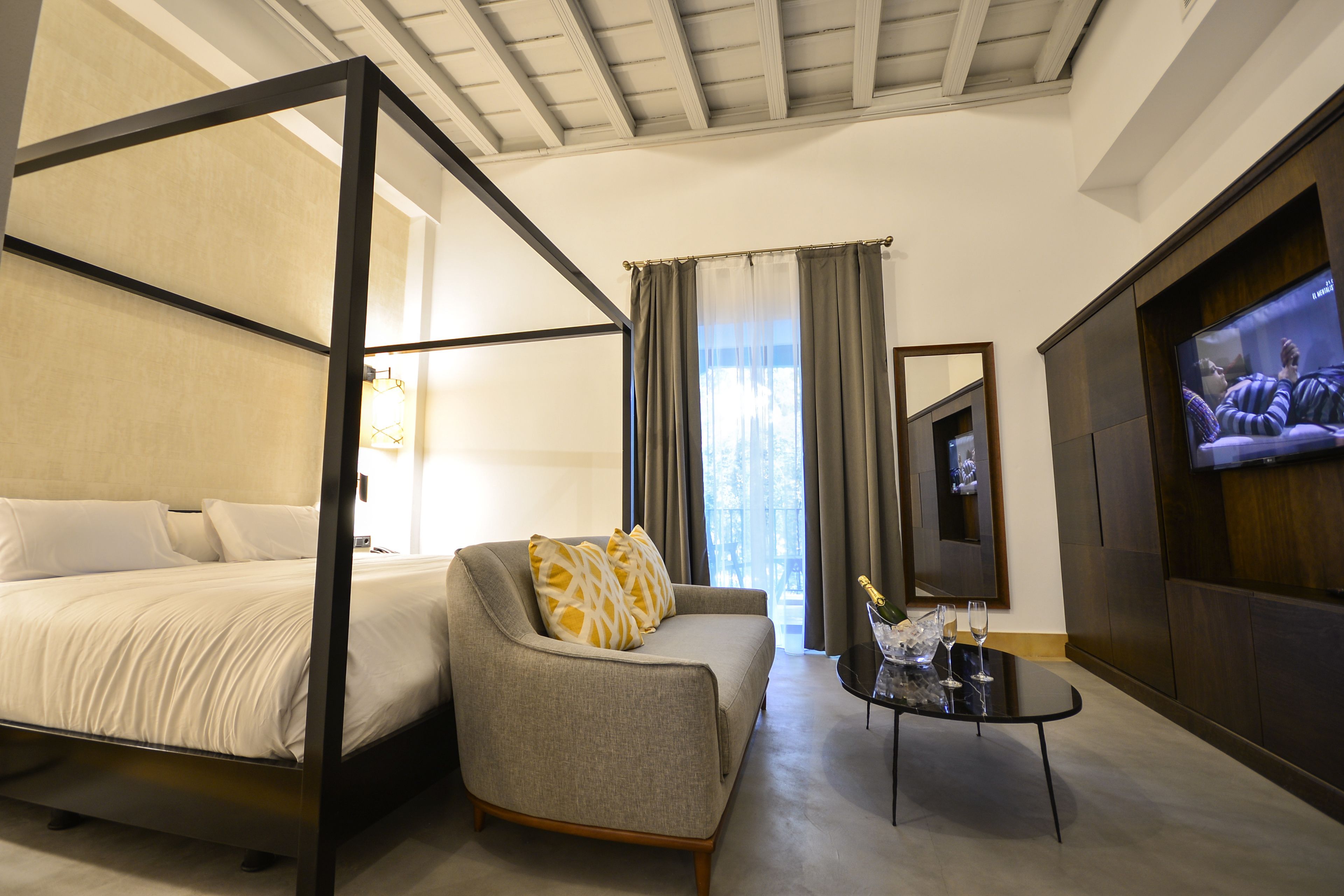 A hotel room with a four-poster bed, a sofa, a TV, and a small table with a bottle and glasses.