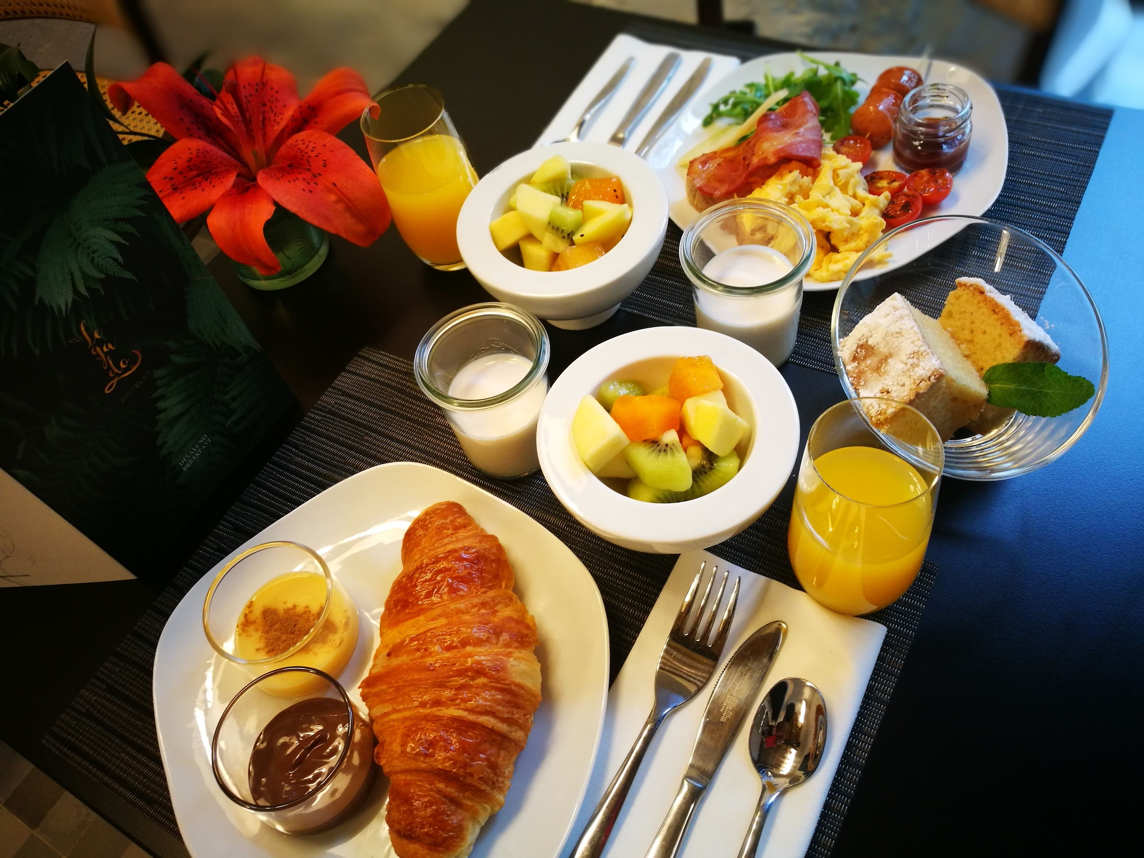 Une table de petit-déjeuner avec des croissants, des fruits, des œufs brouillés, du bacon et un petit gâteau.