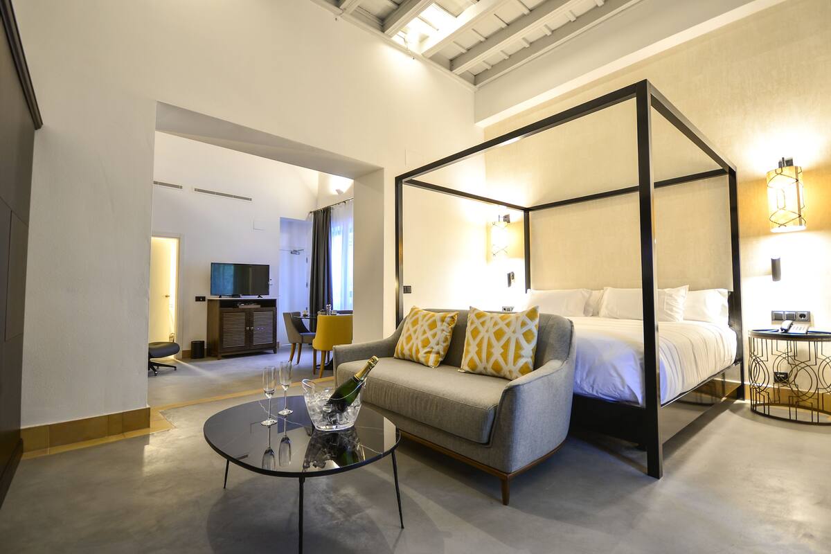 A modern hotel room with a four-poster bed, a sofa, a glass table with a champagne bottle, and a TV in the background.