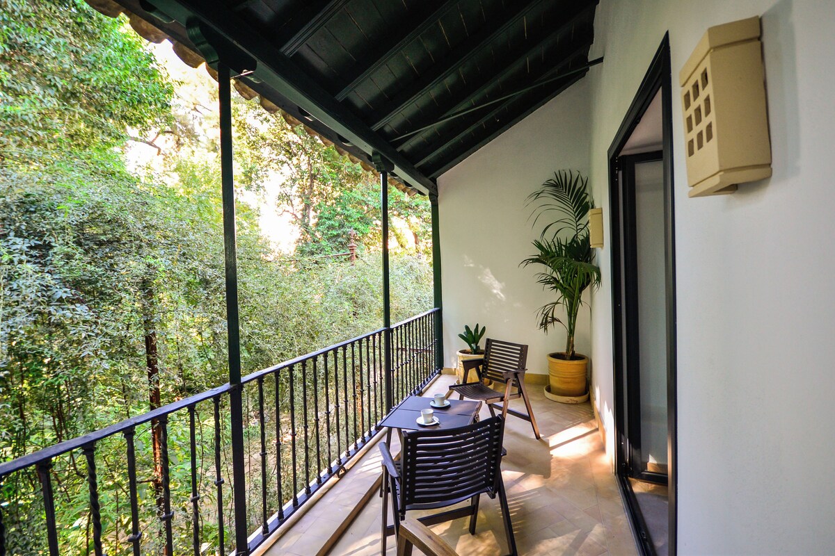 A balcony with a table and chairs, a potted plant, and a view of a lush garden.