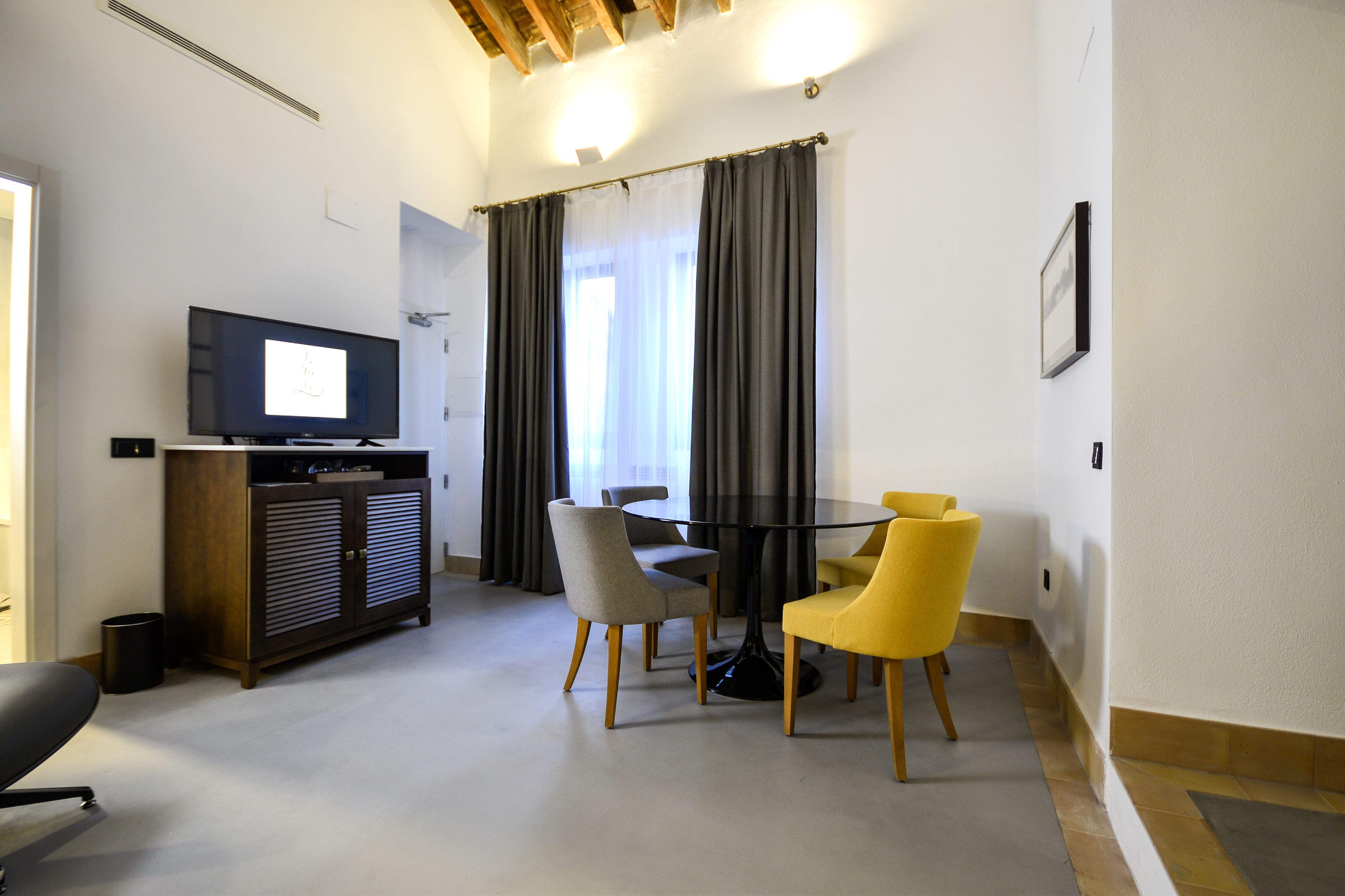 Habitación de hotel con televisión, una mesa de comedor con sillas, un armario y una ventana con cortinas.