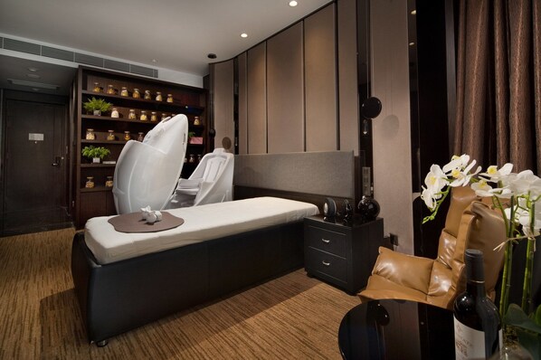 Una habitación de hotel moderna con una cama grande, una mesita de noche con una botella, un mueble pequeño y un estante en la pared con plantas y frascos.