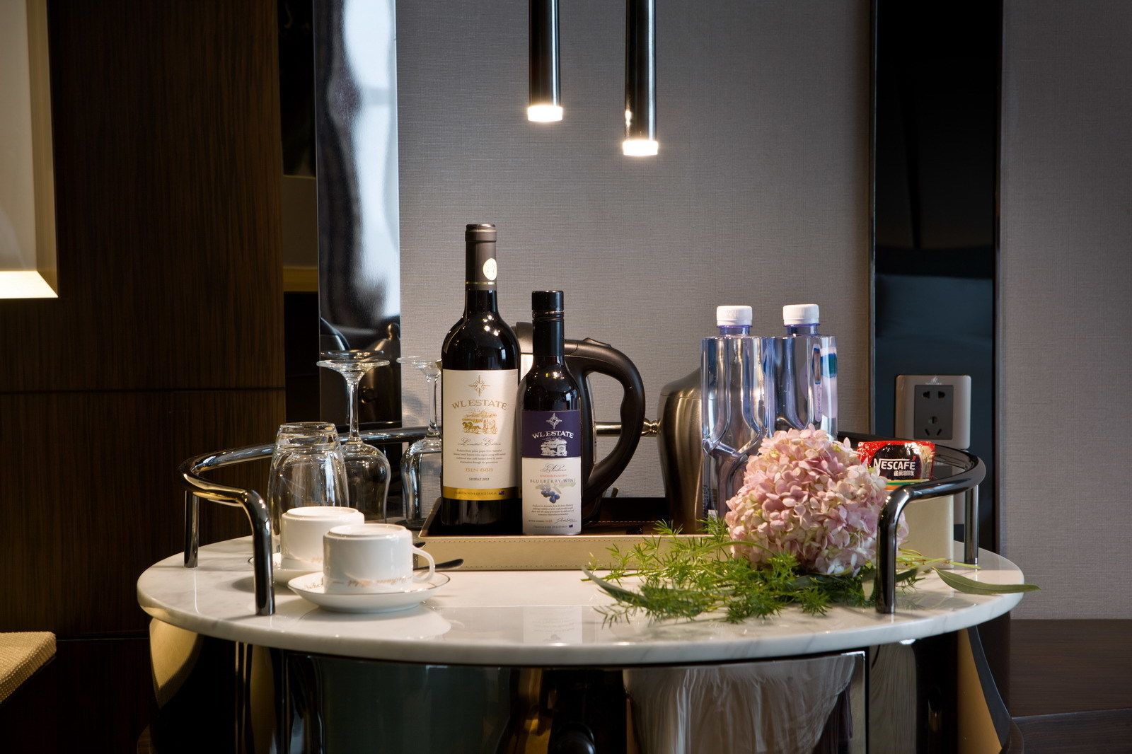 A modern bathroom with a round sink, two bottles of W.L. Estate wine, a Nescafe coffee maker, and a mirror reflecting the room.