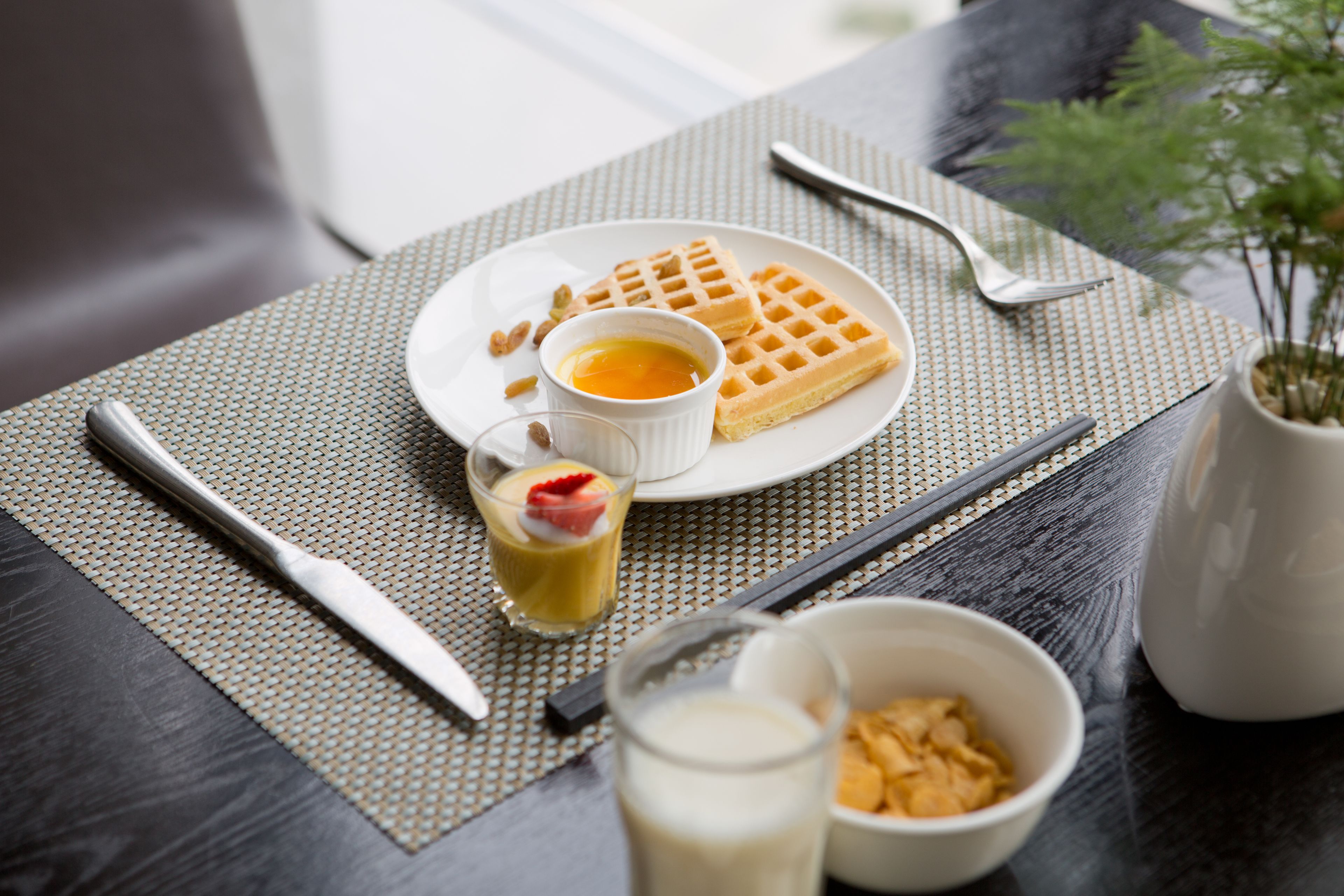 A table with a plate of waffles, a small bowl of fruit, a glass of milk, and a cup of tea.