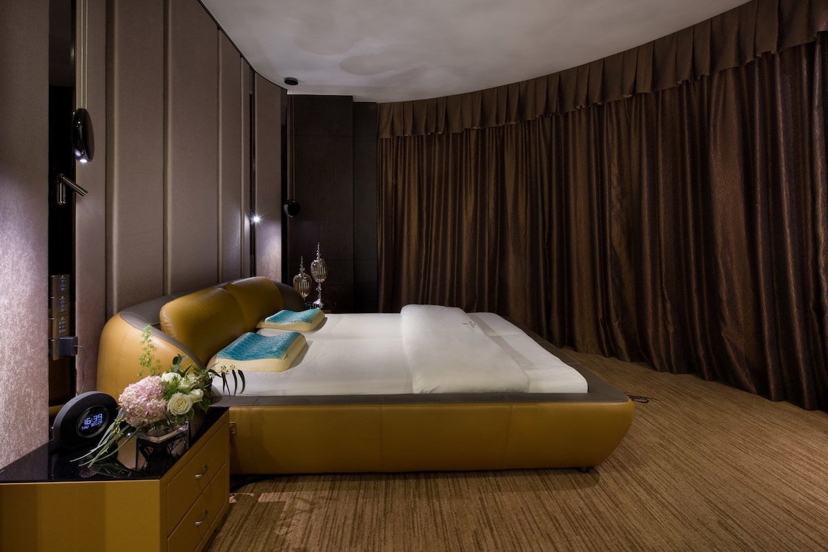 A modern hotel room with a large bed, a bedside table with a clock, a vase with flowers, and a curtain.