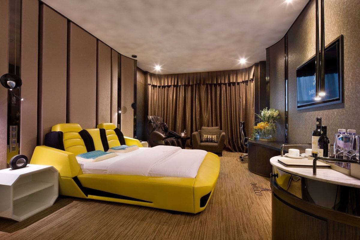 A modern hotel room with a yellow bed, a massage chair, a vanity with toiletries, and a TV on the wall.