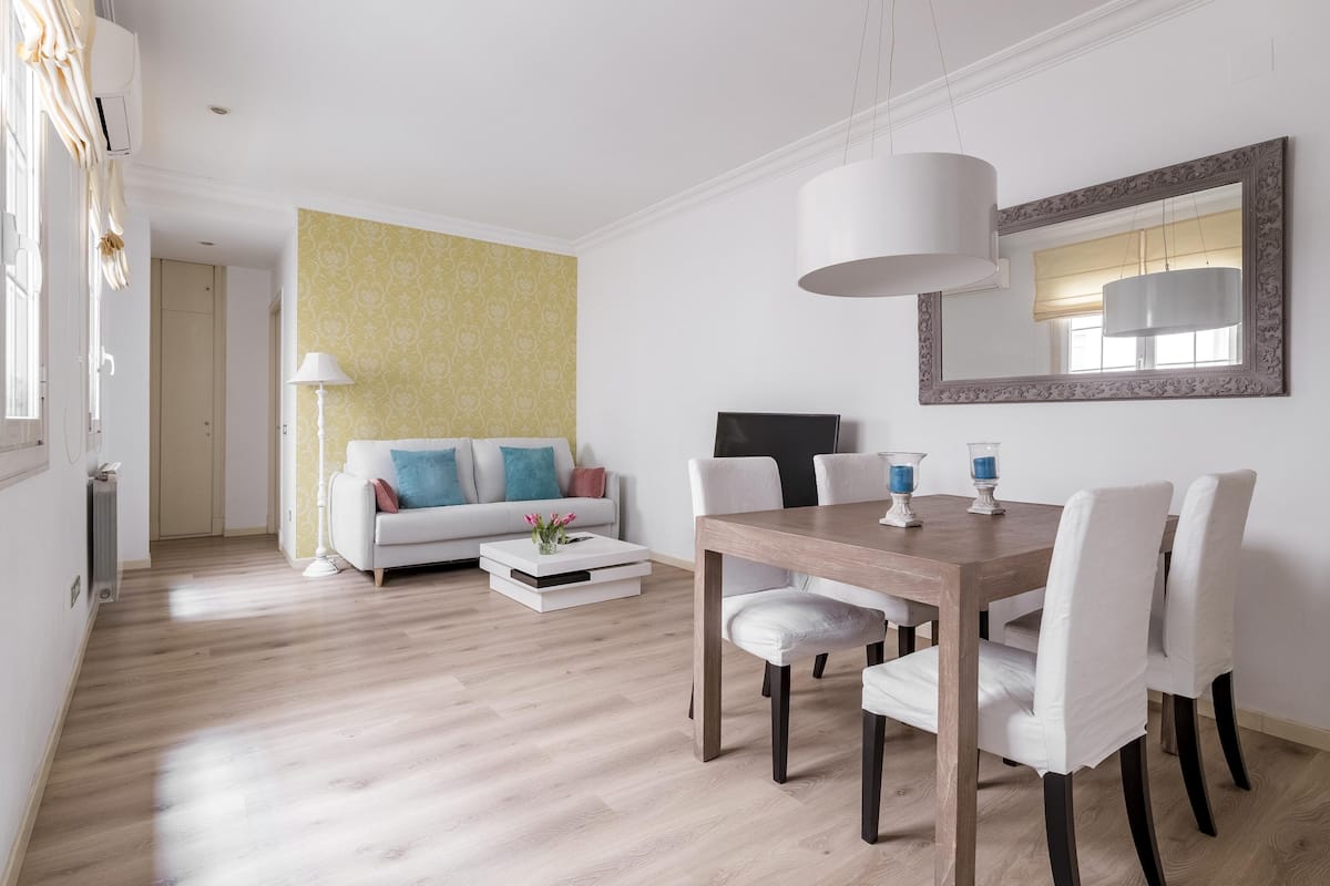 A modern living room with a dining area, featuring a wooden dining table, white upholstered chairs, a sofa with colorful cushions, a patterned wall, and a large mirror.
