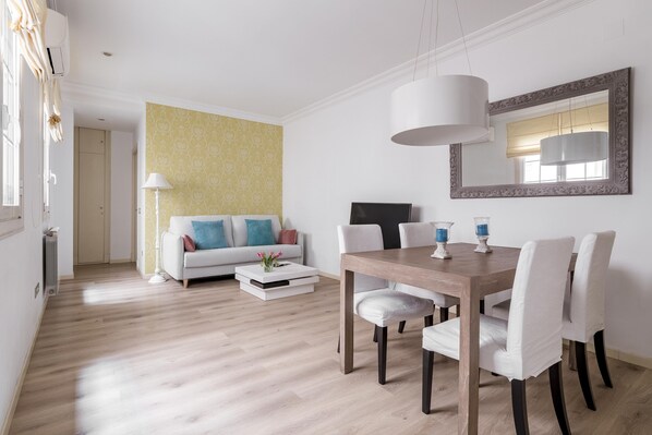 Un salon moderne avec un coin repas, comprenant une table à manger en bois, des chaises à revêtement blanc, un canapé agrémenté de coussins colorés, un mur à motifs et un grand miroir.