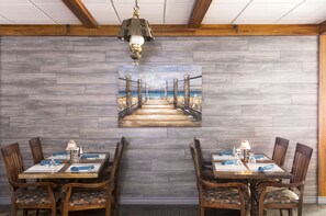 A dining area with a wooden table and chairs, a painting of a pier, and a hanging light fixture. - Mount Peyton Resort and Conference Centre (Grand Falls - Windsor)
